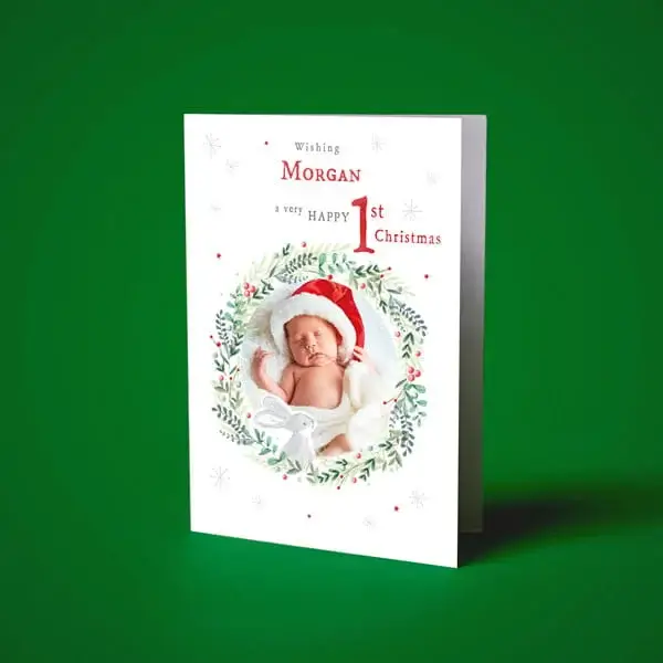 Personalised baby's first Christmas card featuring a newborn photo framed by festive foliage and a red Santa hat design.